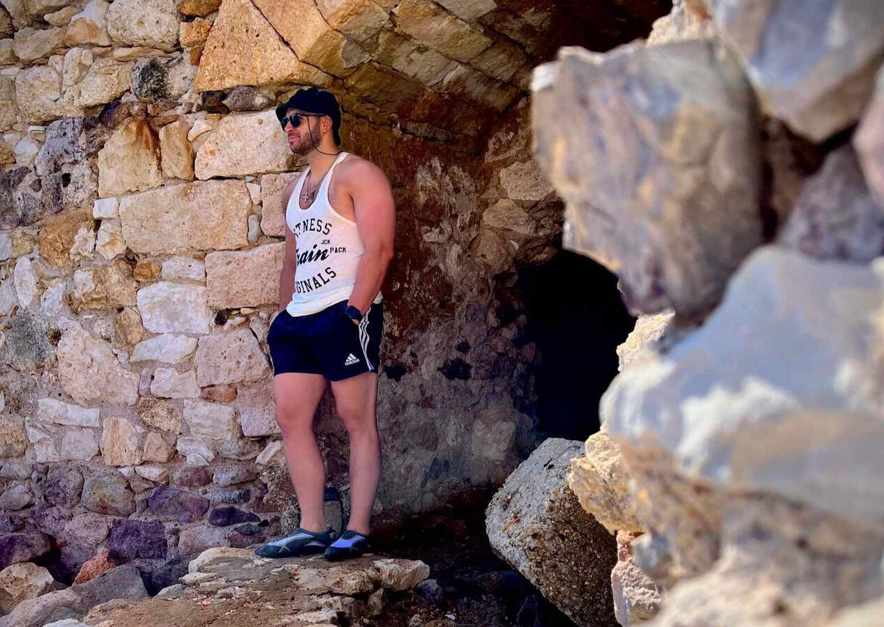 Solo destination photo shoot of man standing on rocks, wearing sunglasses, a hat, shirt and shorts with big rocks in the background in Milos Greece, photo taken by Mia's Photography and Events.