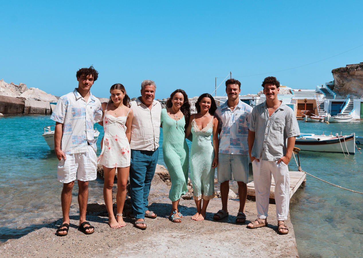 Family photoshoot at Mandrakia in Milos, Greece. The family consists of smiling men and women posing for a family photograph with turquoise water and colourful boat houses in the background