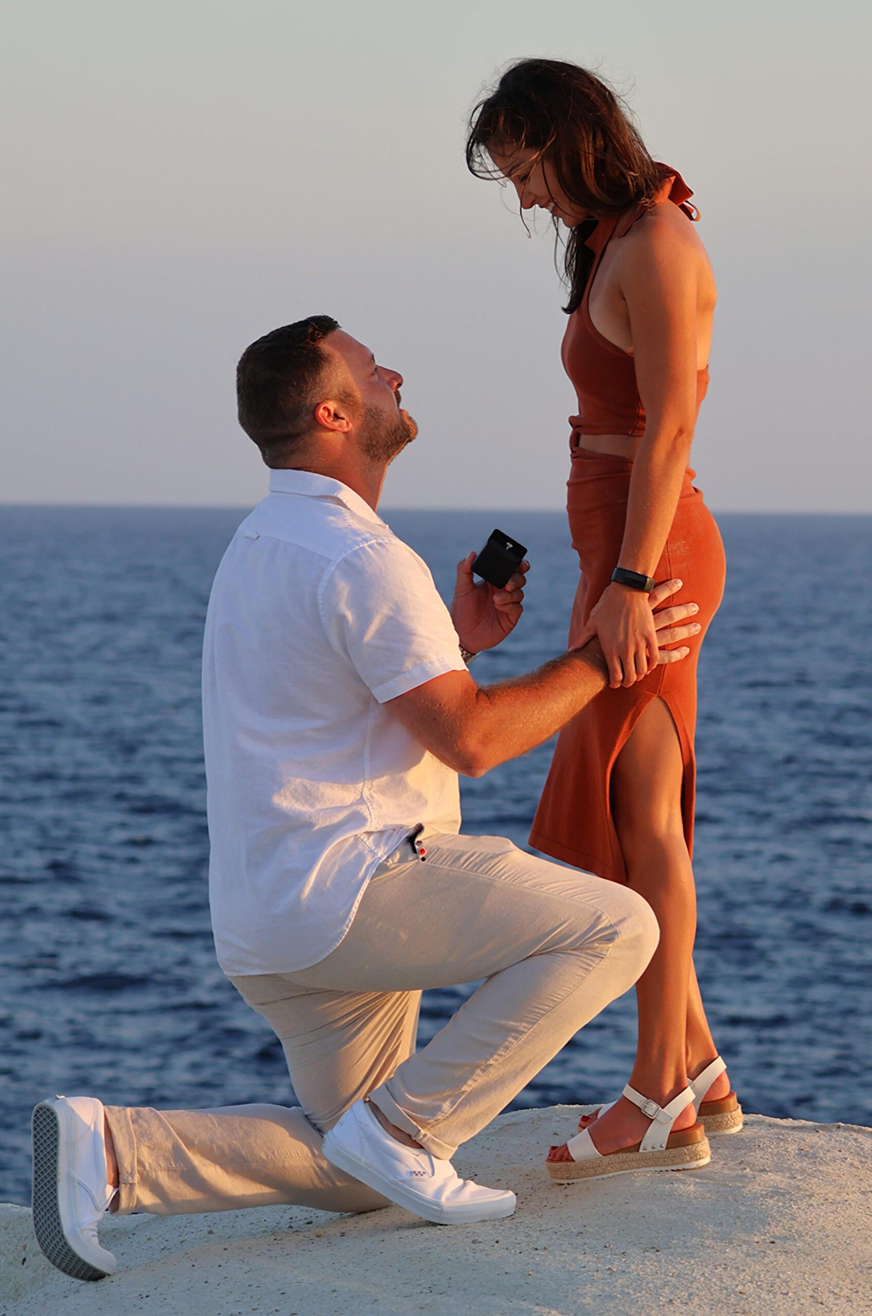 Sunrise at Sarakiniko in Milos Greece, photo taken by Mia's Photography and Events. Person kneeling on one knee, on the white volcanic rocks with a ring in his hand proposing to a lady in a brownish orange dress