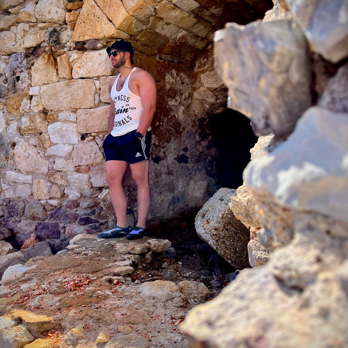 Solo destination photo shoot of man standing on beach, wearing sunglasses, a black hat, white shirt and black shorts with sea and stones in the background in Milos Greece, photo taken by Mia's Photography and Events.