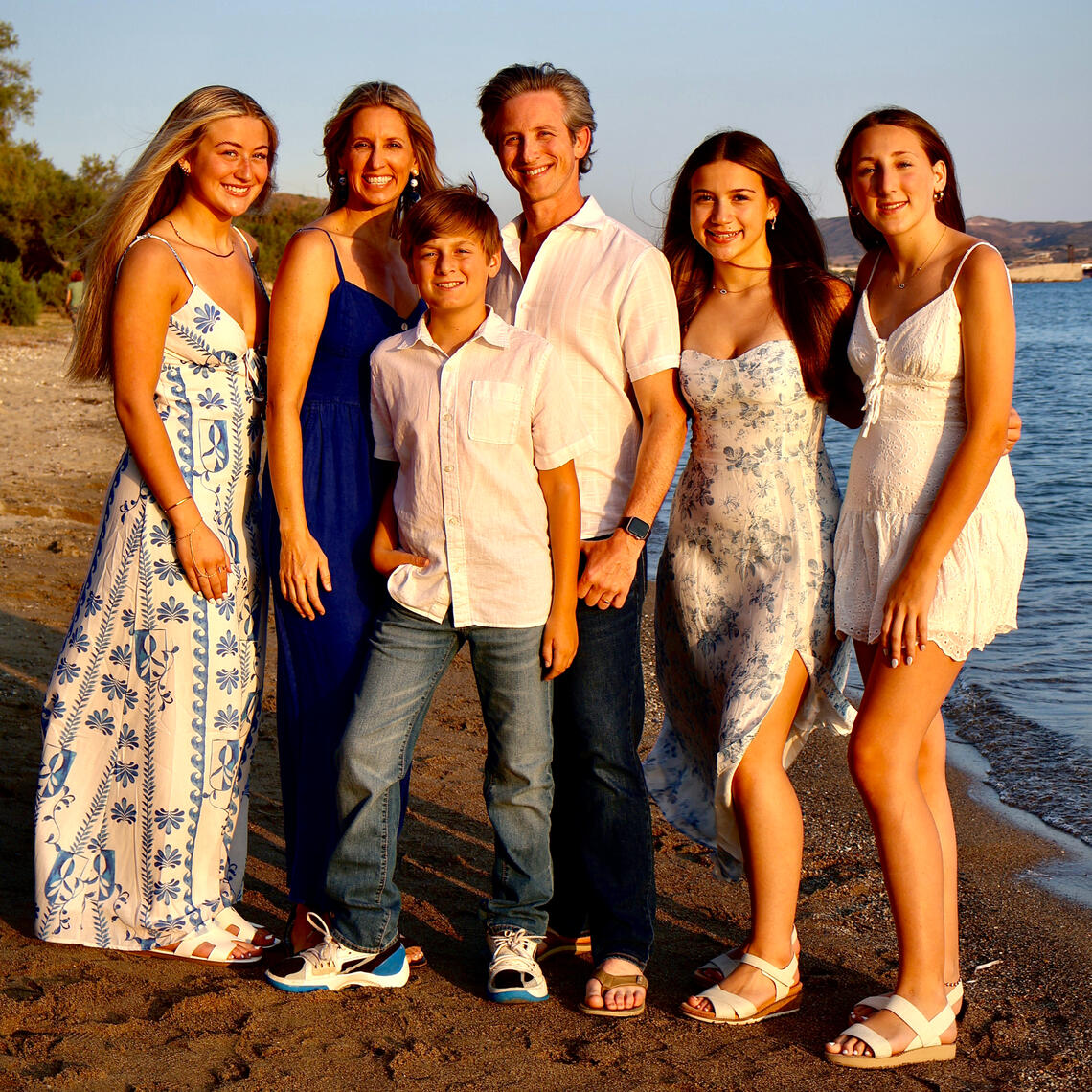 Family of 6 having a photoshoot on a sandy beach in Milos with the blue sea on the right hand side. Everyone is smiling and there is a beautiful golden hour light as this is at sunset time.