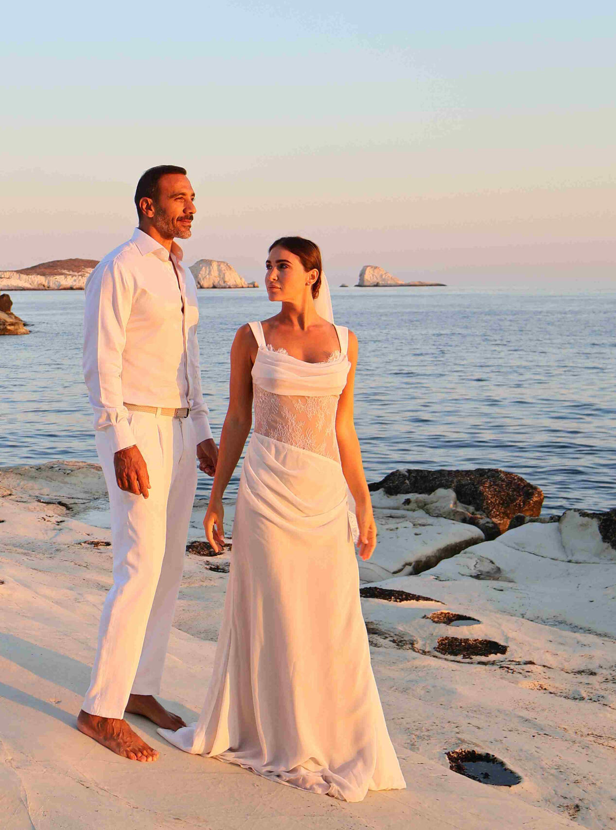 Solo destination photo shoot of man standing on rocks, wearing sunglasses, a hat, shirt and shorts with big rocks in the background in Milos Greece, photo taken by Mia's Photography and Events.